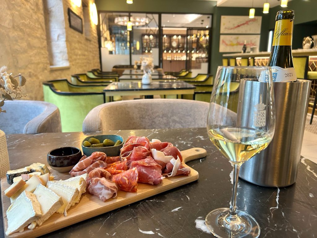 A board of charcuterie and cheese beside a glass of white wine and a bottle of Olivier Leflaive 2021 in an ice bucket