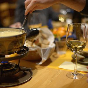 A fondue pot over a small burner on a wooden table, a long fork in the cheese and a glass of white wine beside it