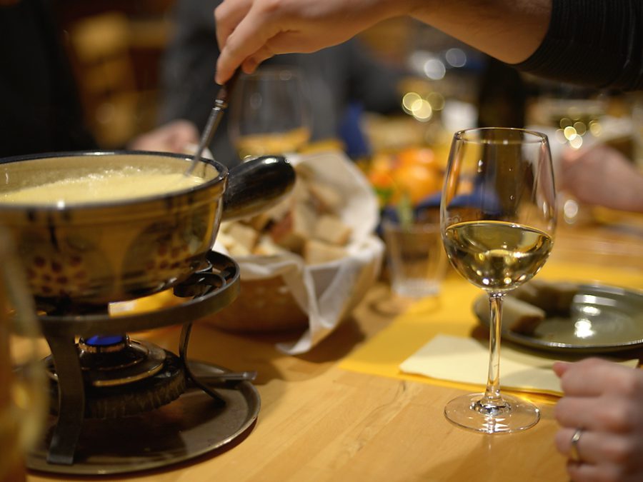 A fondue pot over a small burner on a wooden table, a long fork in the cheese and a glass of white wine beside it