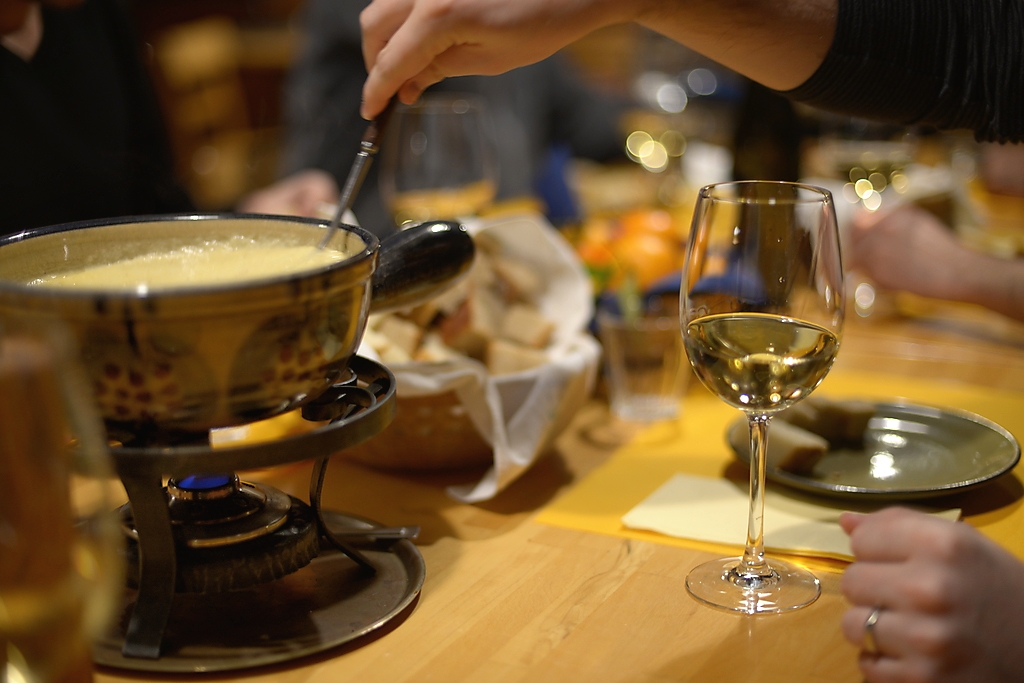 A fondue pot over a small burner on a wooden table, a long fork in the cheese and a glass of white wine beside it