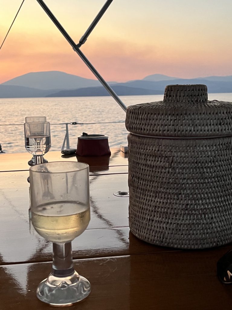 A glass of white wine and a woven ice bucket on a yacht's wooden table at sunset, hills across the water